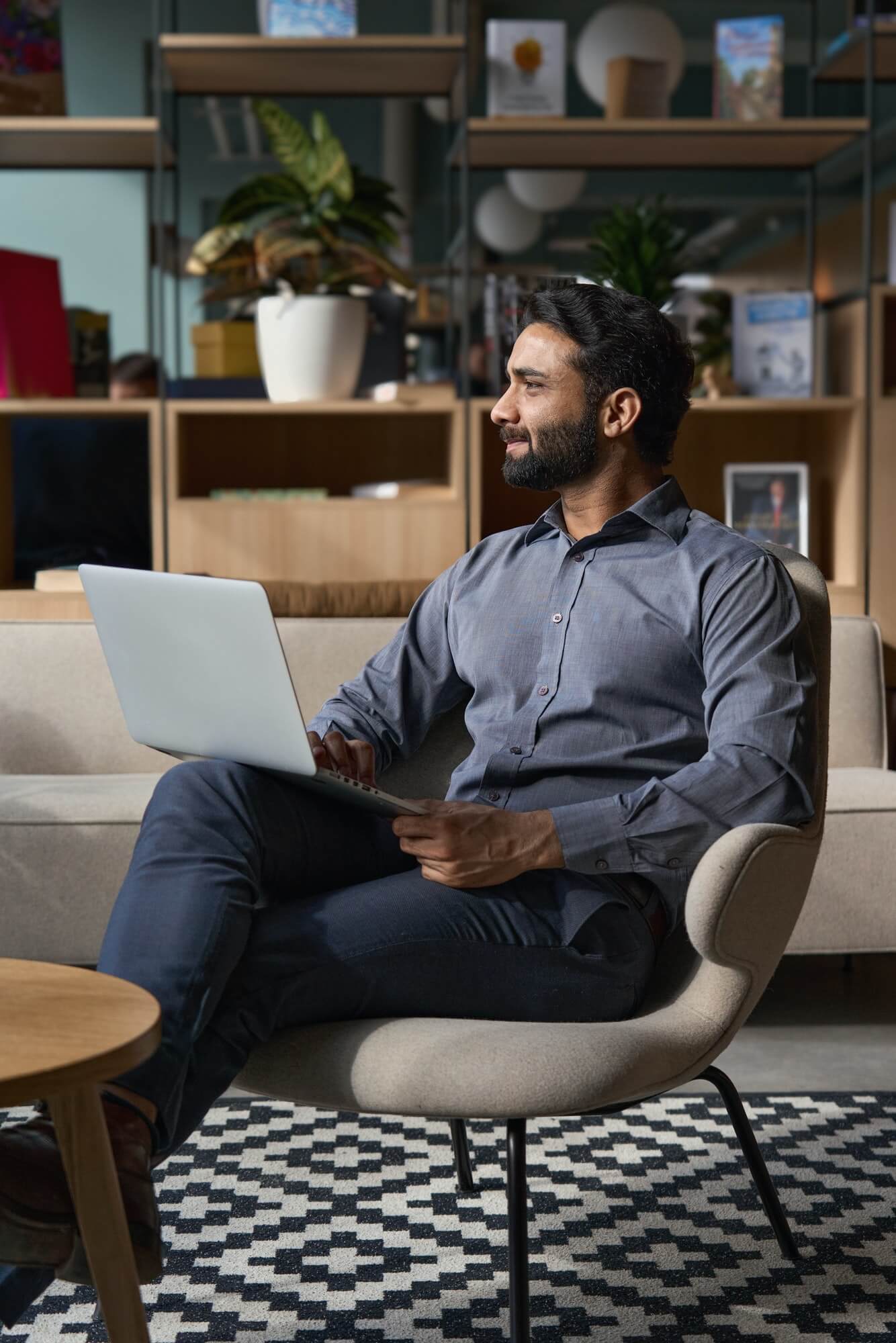 Indian business man looking away sitting on chair in modern office using laptop.